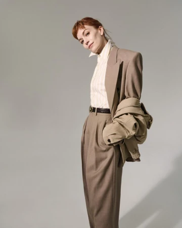 A woman in a tan suit poses in front of a neutral background, with a jacket draped over her shoulder in a studio fashion shot.