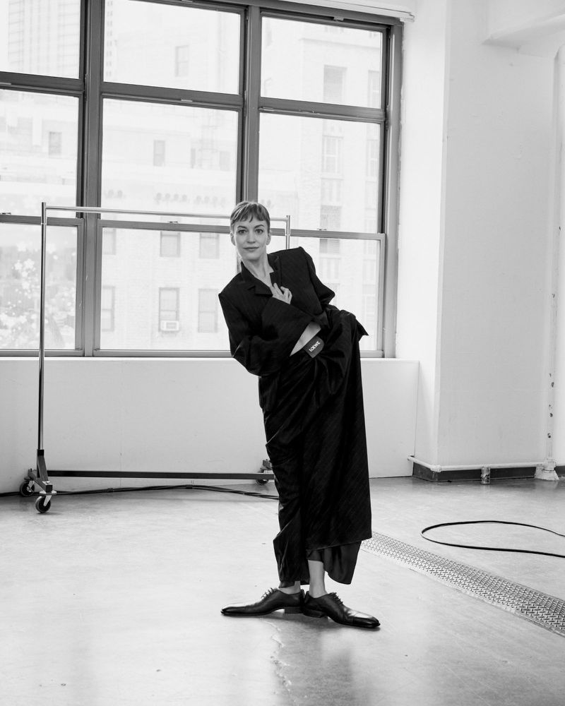 A woman in a black jacket and skirt poses indoors near a window with buildings visible outside in a black and white fashion shot.