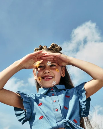 A young girl with a denim shirt and flower stickers on her face shields her eyes while looking up at the blue sky with clouds.