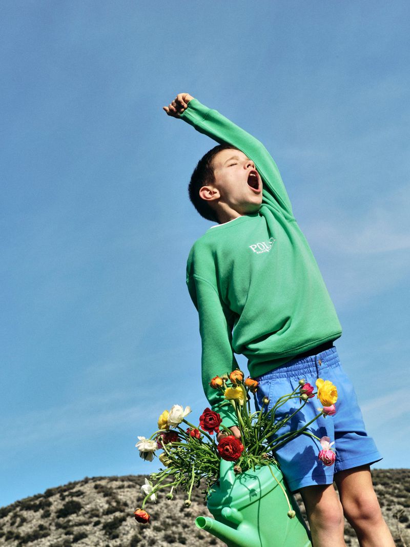 A joyful young boy stands with a green watering can filled with flowers, set against a clear blue sky and rolling hills. 