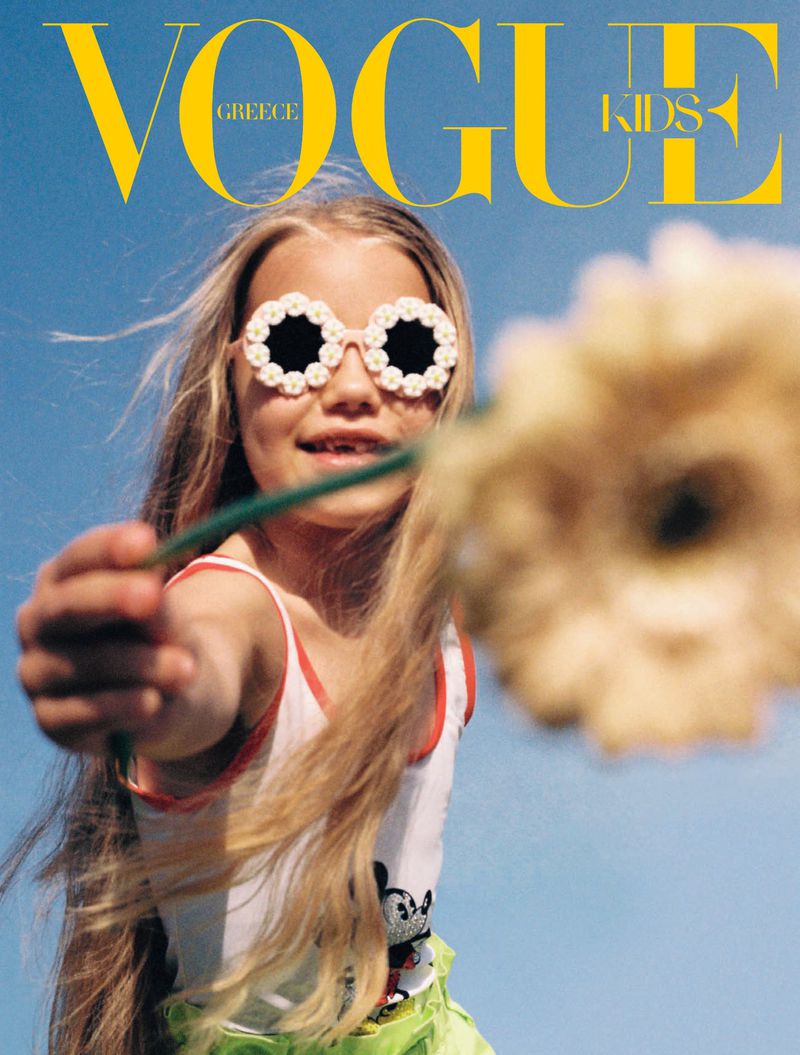 A young blonde child with flower-shaped sunglasses holds a flower in front of a blue sky for the cover of Vogue Greece Kids.