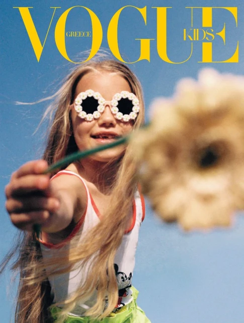 A young blonde child with flower-shaped sunglasses holds a flower in front of a blue sky for the cover of Vogue Greece Kids.