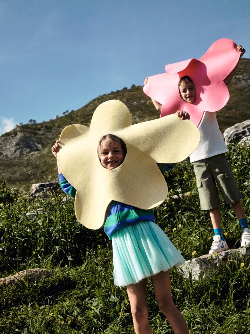 Two children in flower costumes pose outdoors on a sunny day. One wears a yellow flower, the other a pink one, smiling in a field.