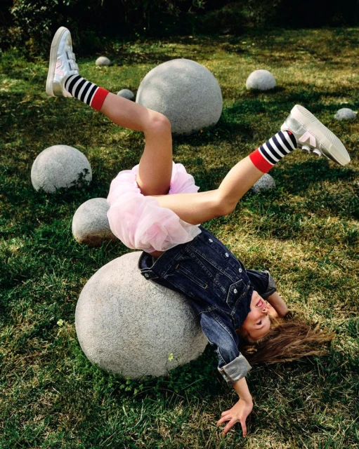 Child upside down, resting on a granite ball in a yard. The child is wearing a pink tutu, jean jacket, striped socks and sneakers.