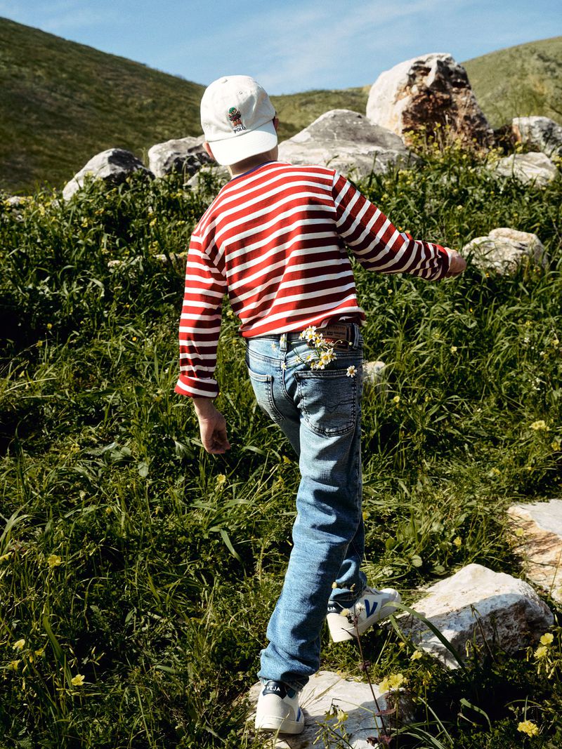 A child in jeans and a striped shirt walking through a grassy field with scattered rocks, wearing a hat, enjoying a sunny day outdoors.