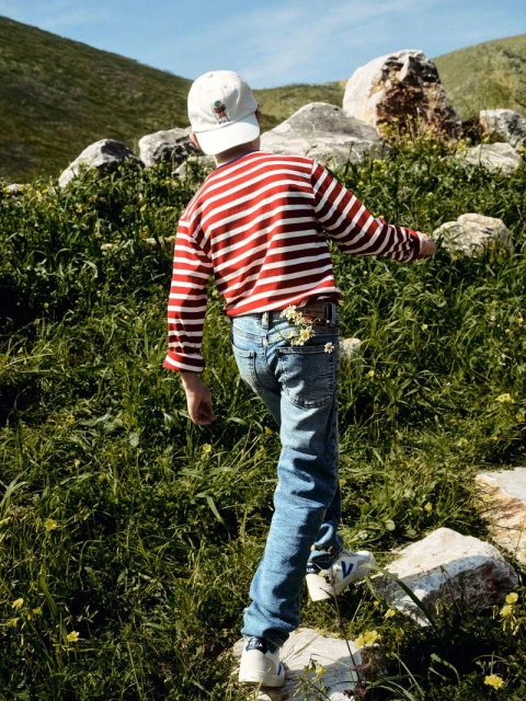 A child in jeans and a striped shirt walking through a grassy field with scattered rocks, wearing a hat, enjoying a sunny day outdoors.