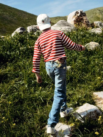 A child in jeans and a striped shirt walking through a grassy field with scattered rocks, wearing a hat, enjoying a sunny day outdoors.