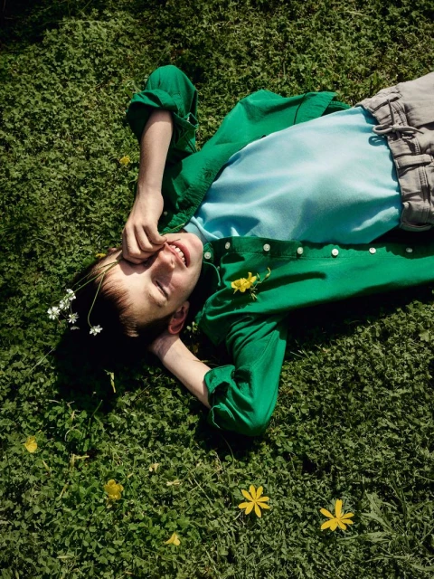 Child in green cardigan lies on a sunny lawn covered with grass and small yellow and white wildflowers, looking up and smiling.