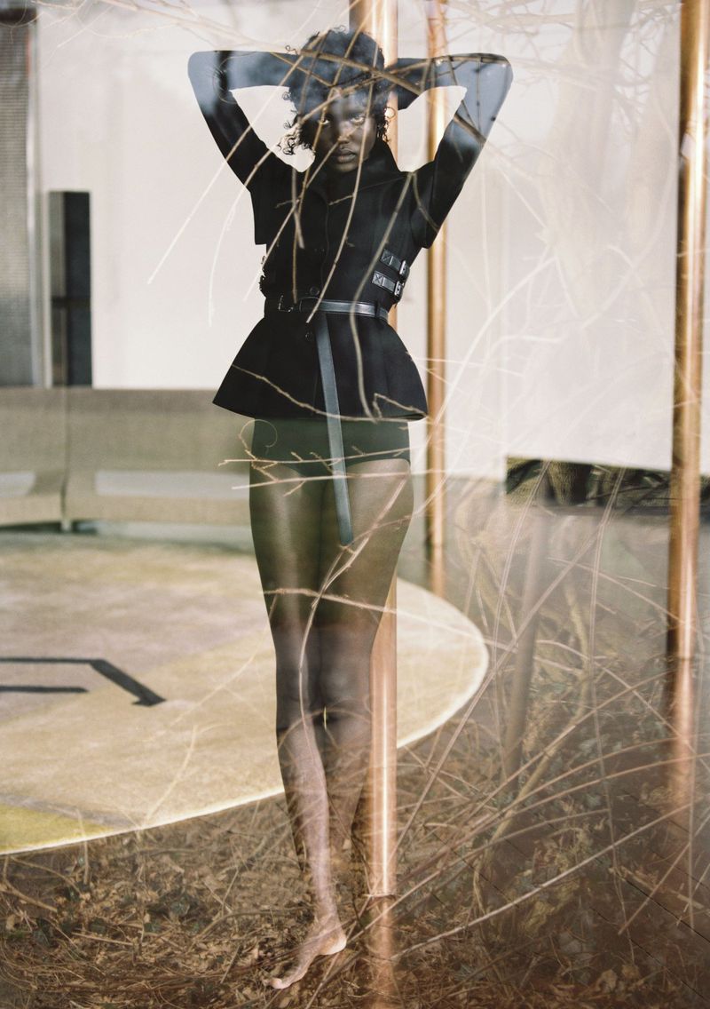 Adult female fashion model strikes a full-length pose indoors wearing a black jacket with a belt and high-waisted bottoms, plus decorative twigs.