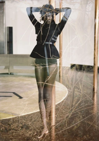 Adult female fashion model strikes a full-length pose indoors wearing a black jacket with a belt and high-waisted bottoms, plus decorative twigs.