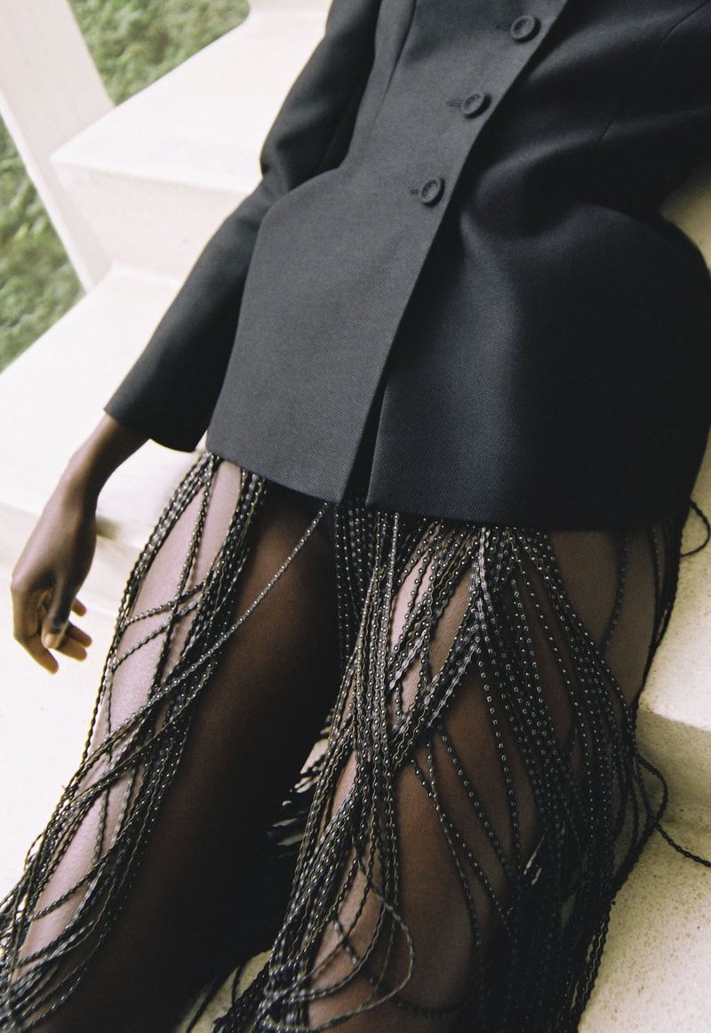 A fashion shot features a dark-skinned model wearing a black blazer and beaded fringe pants leaning against a white concrete staircase.