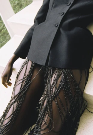 A fashion shot features a dark-skinned model wearing a black blazer and beaded fringe pants leaning against a white concrete staircase.