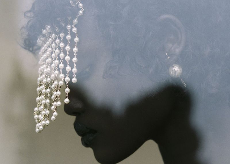 Portrait of a dark-skinned woman wearing a pearl headpiece, featuring a delicate earring and makeup, against a neutral background.