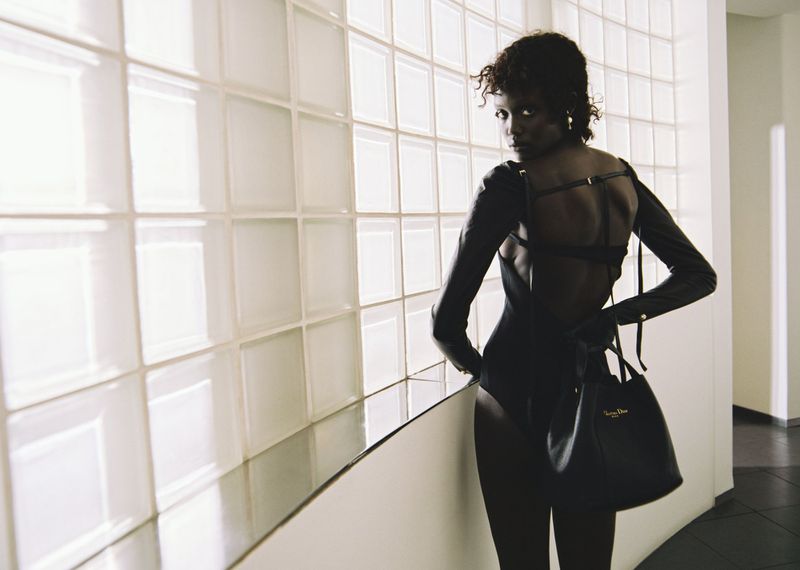 An adult female model with a dark complexion poses, glancing back over her shoulder, wearing a black bodysuit and holding a black handbag.