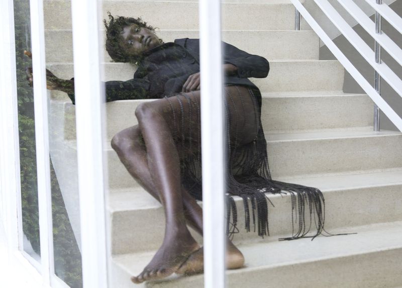 Fashion portrait of a Black model reclining on stairs wearing a black jacket and fringed skirt, with a glimpse of greenery in the background.