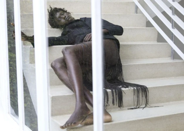 Fashion portrait of a Black model reclining on stairs wearing a black jacket and fringed skirt, with a glimpse of greenery in the background.