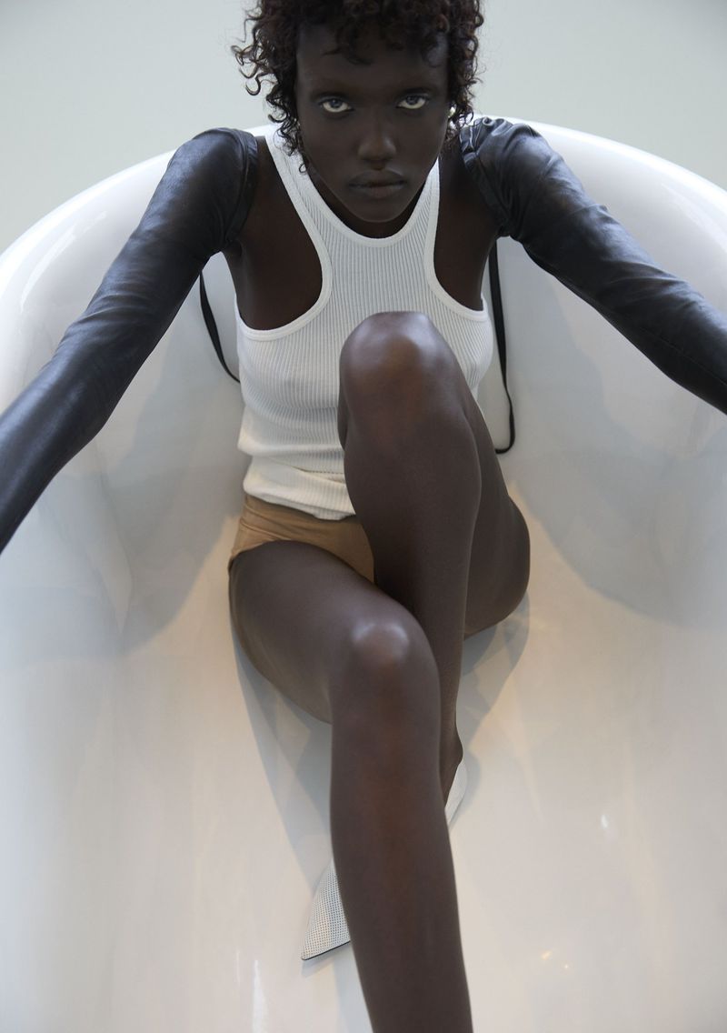 Adult female model poses in a white bathtub with black arm coverings, wearing a white ribbed tank top and tan shorts, in studio lighting.