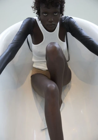 Adult female model poses in a white bathtub with black arm coverings, wearing a white ribbed tank top and tan shorts, in studio lighting.