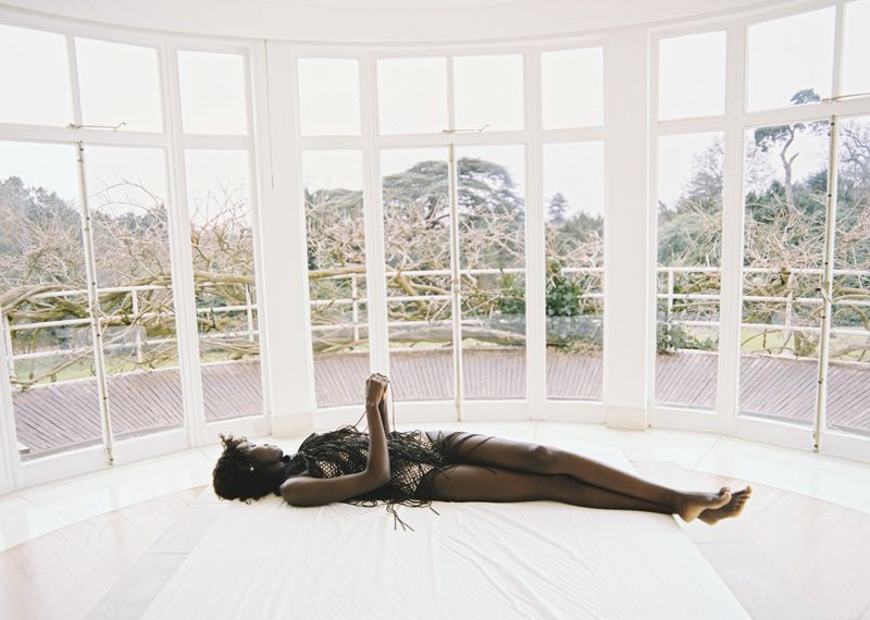 A woman in a dark, fringed garment lies on a white surface in a bright room with large windows, looking out at trees.
