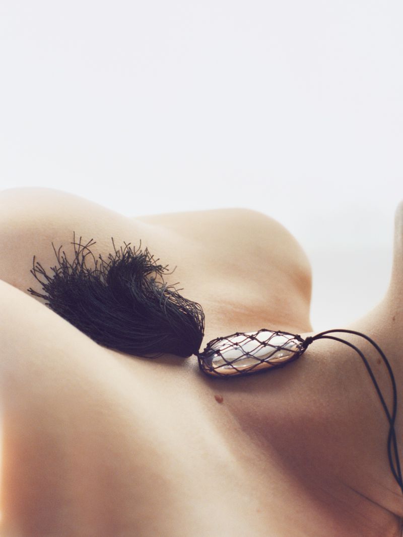 Nude torso of an adult female with a black tassel and pendant on a simple white background, showcasing soft diffused lighting.