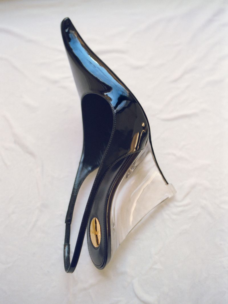 Close-up studio shot of a glossy black patent leather slingback wedge heel shoe with a clear wedge heel on a white surface.