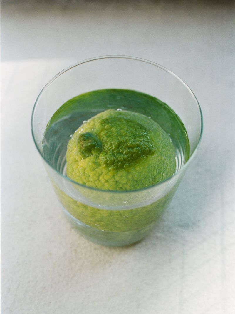 Still life shot of a textured green citrus fruit submerged in a clear glass filled with water, set against a bright white background.