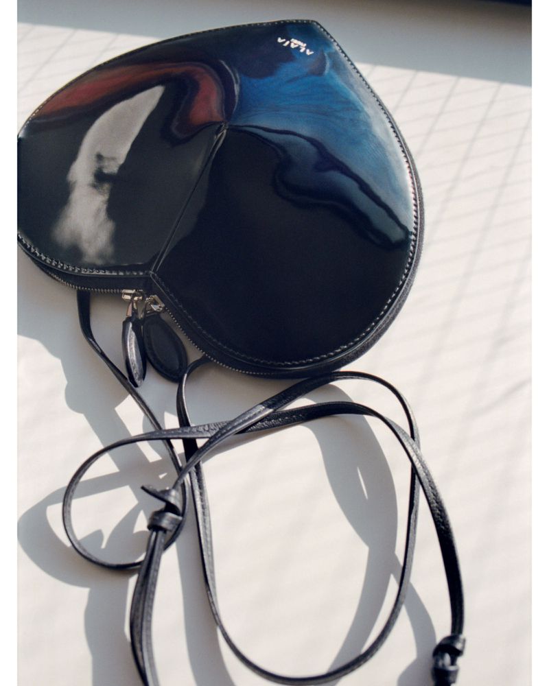 Heart-shaped black patent leather handbag with a crossbody strap. The bag features a zipper closure and a designer logo is visible.