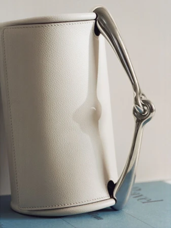 Elegant ivory-colored leather handbag featuring a distinctive metal handle, showcased against a soft, neutral backdrop.