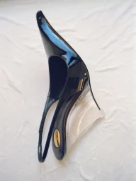 Close-up product shot of a black patent leather slingback shoe with a clear transparent wedge heel against a white fabric background.