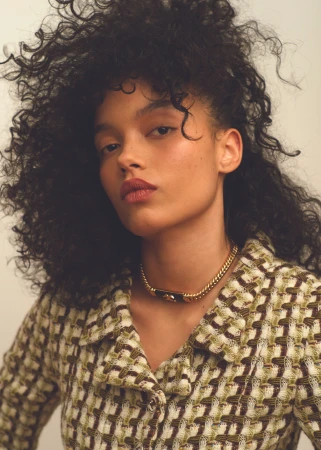 Portrait of Whitney Peak wearing a patterned tweed jacket and gold chain necklace against a light background