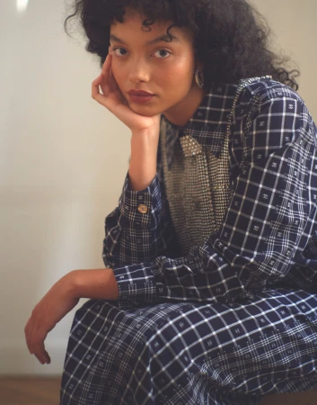 Whitney Peak wearing a blue plaid collared shirt and skirt with her chin resting in her hand against a neutral background.