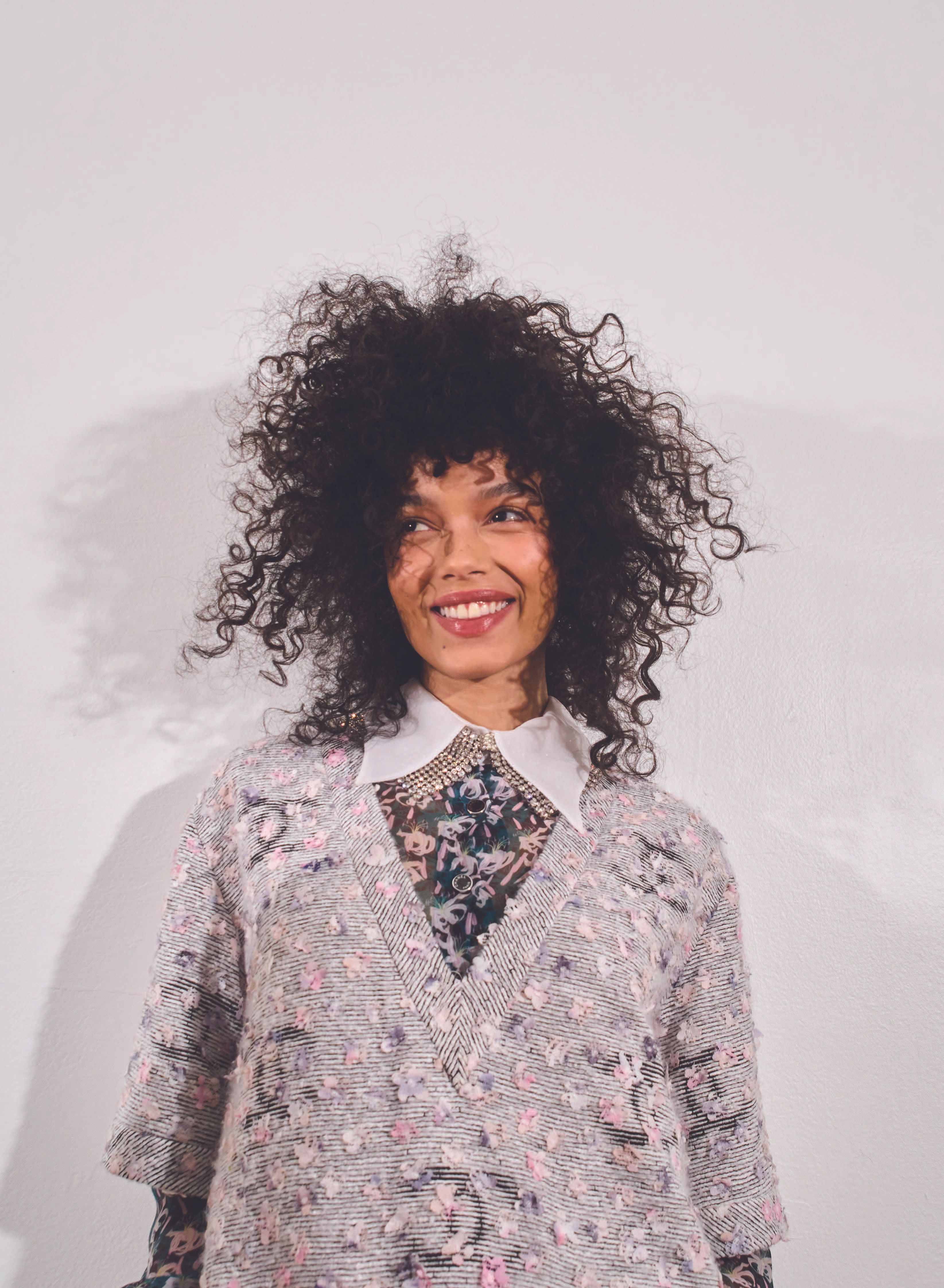 Smiling woman with curly hair wearing a patterned sweater with floral accents and a white collared shirt against a white backdrop.
