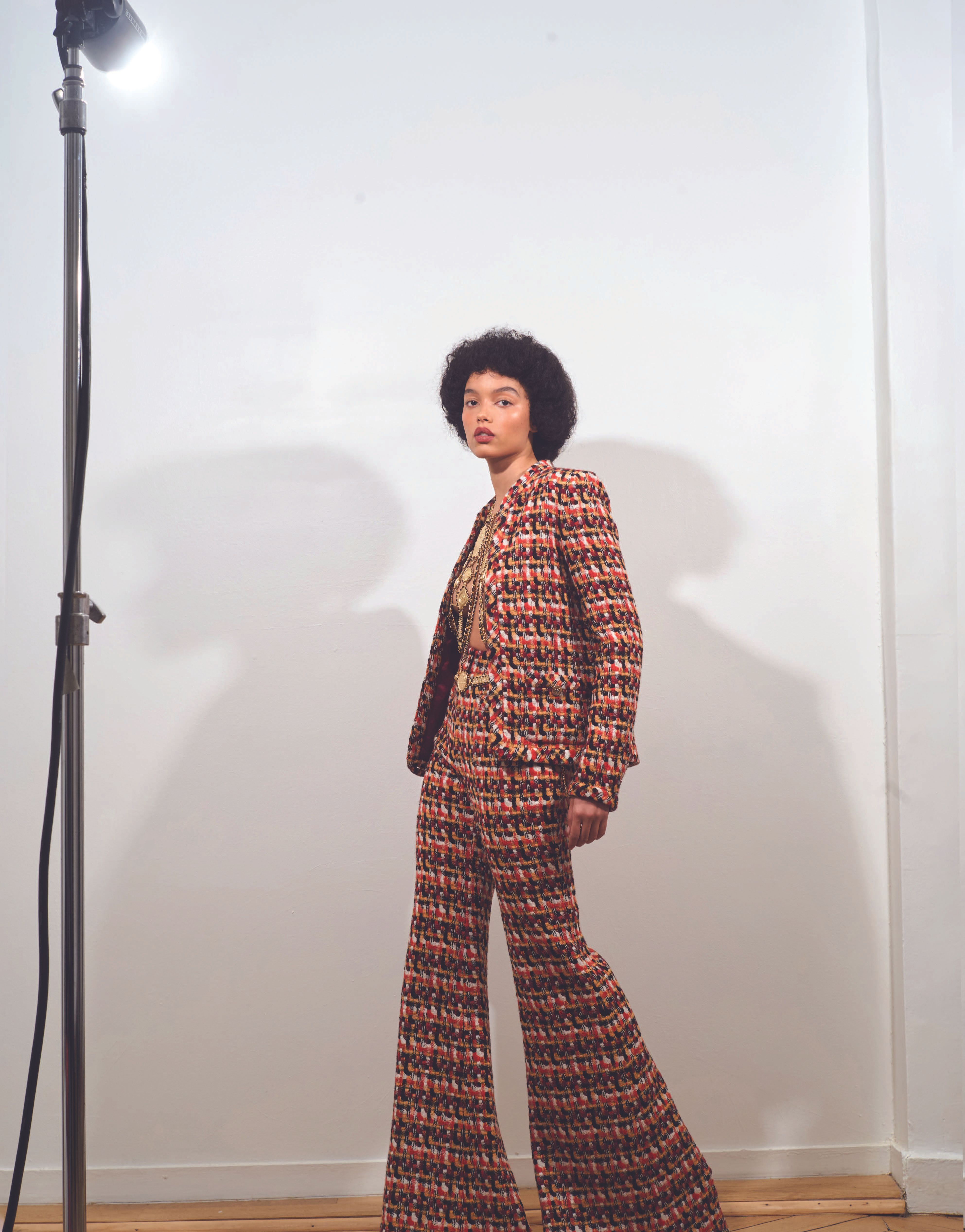A woman in a patterned pantsuit stands in a studio setting with a light stand, posing for a fashion editorial shoot.