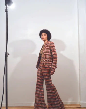A woman in a patterned pantsuit stands in a studio setting with a light stand, posing for a fashion editorial shoot.
