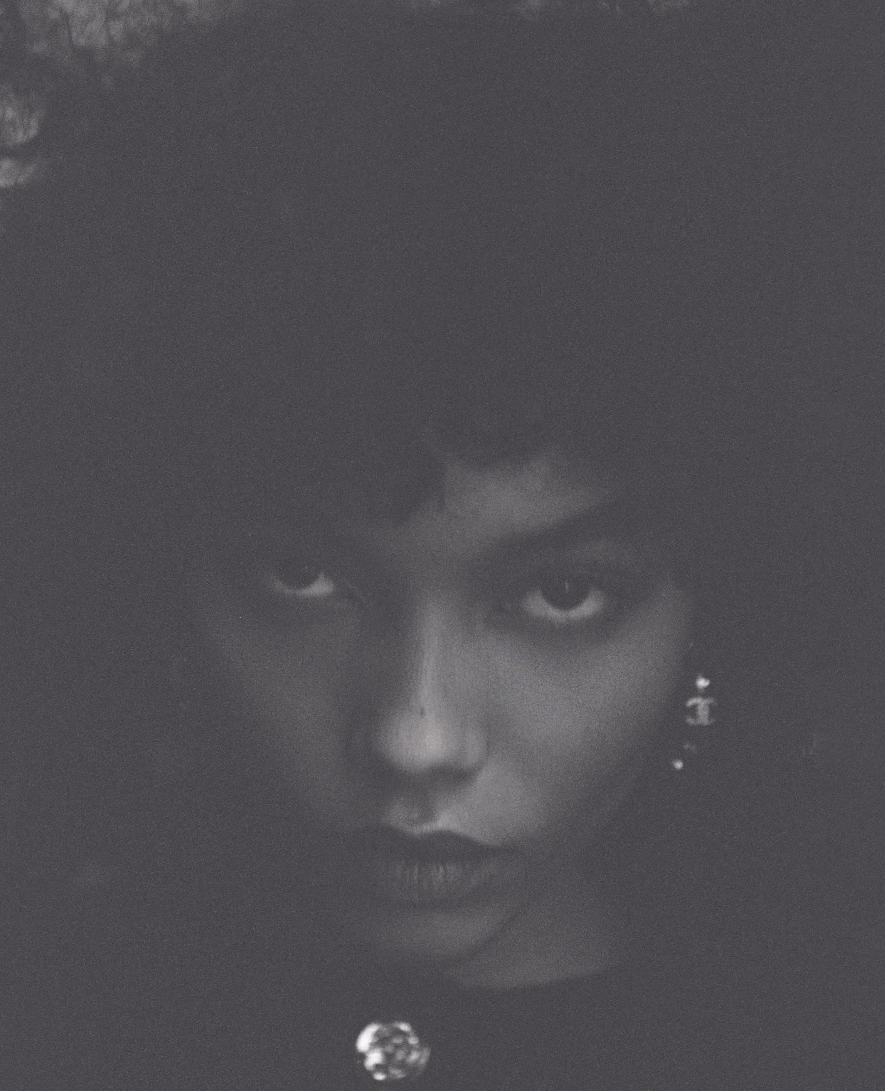 Monochrome portrait of a young woman with an afro hairstyle, featuring close-up framing and elegant jewellery, shot in subdued lighting.