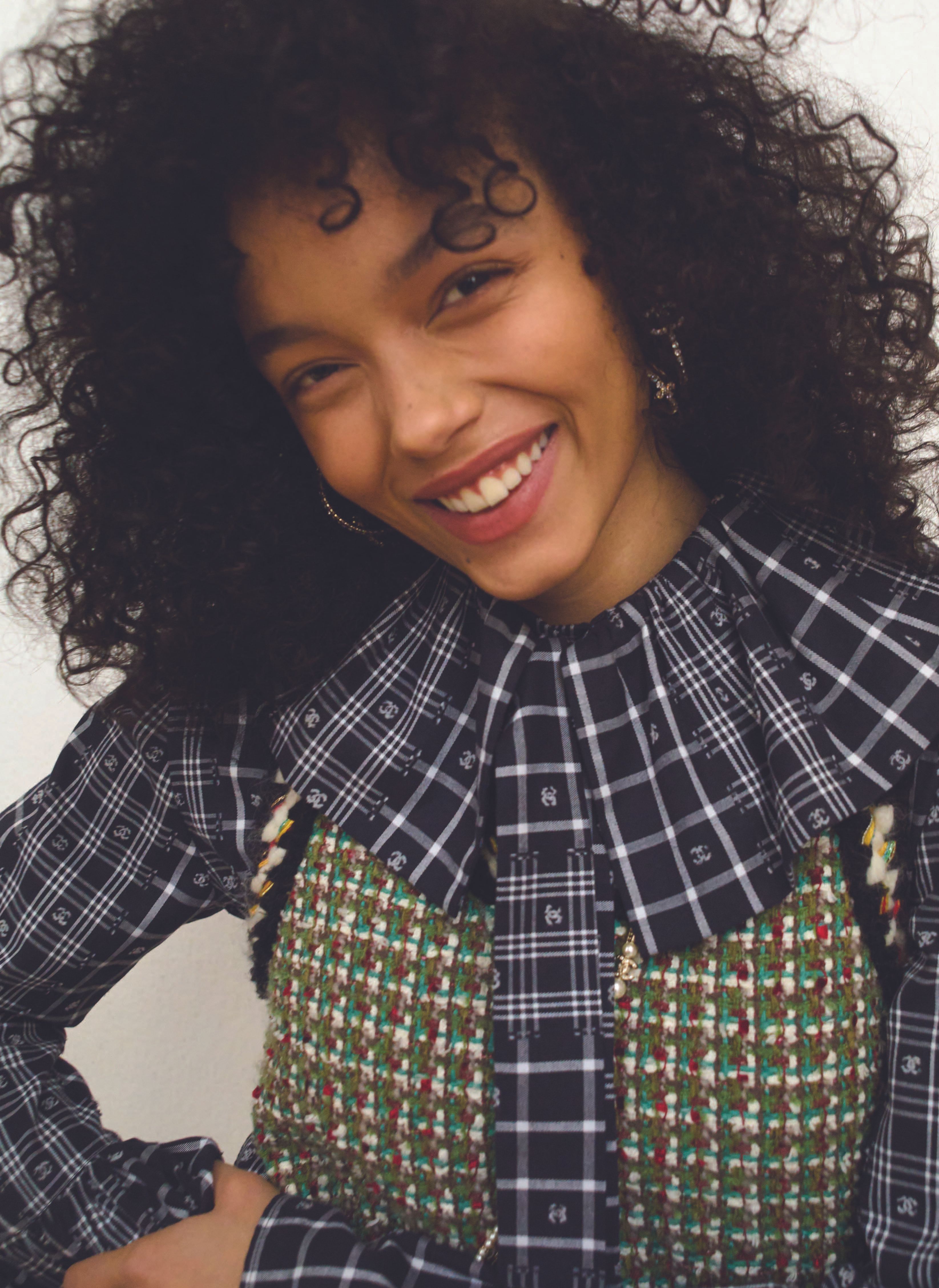 Close-up of a cheerful woman with curly hair smiling, wearing a plaid collared shirt and tweed top against a neutral backdrop.