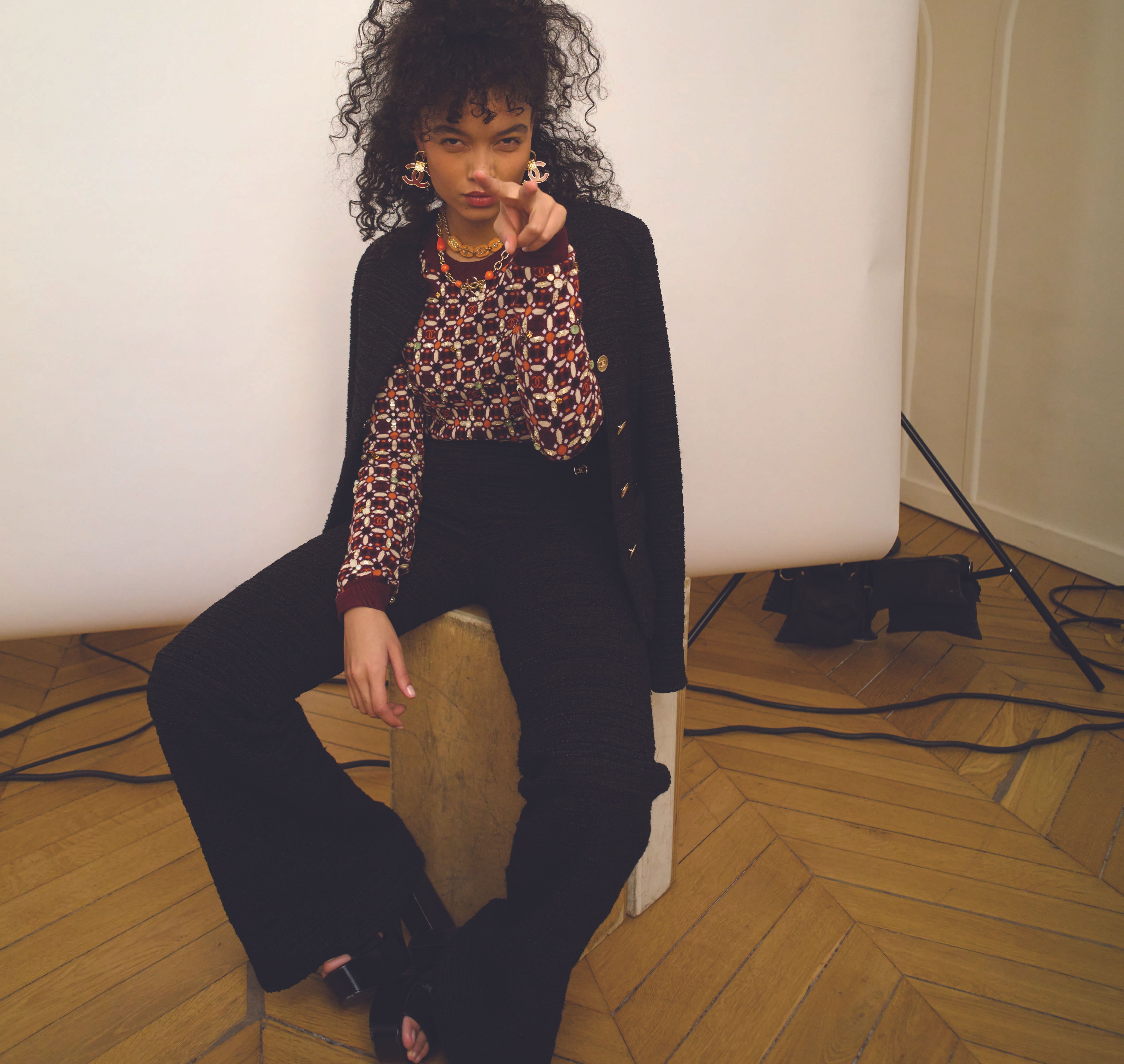 A young woman with curly hair poses indoors, sitting and pointing her finger at the camera, wearing a patterned top and black pants.
