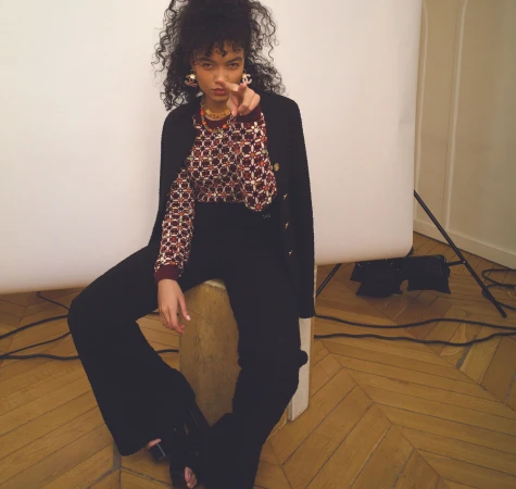 A young woman with curly hair poses indoors, sitting and pointing her finger at the camera, wearing a patterned top and black pants.
