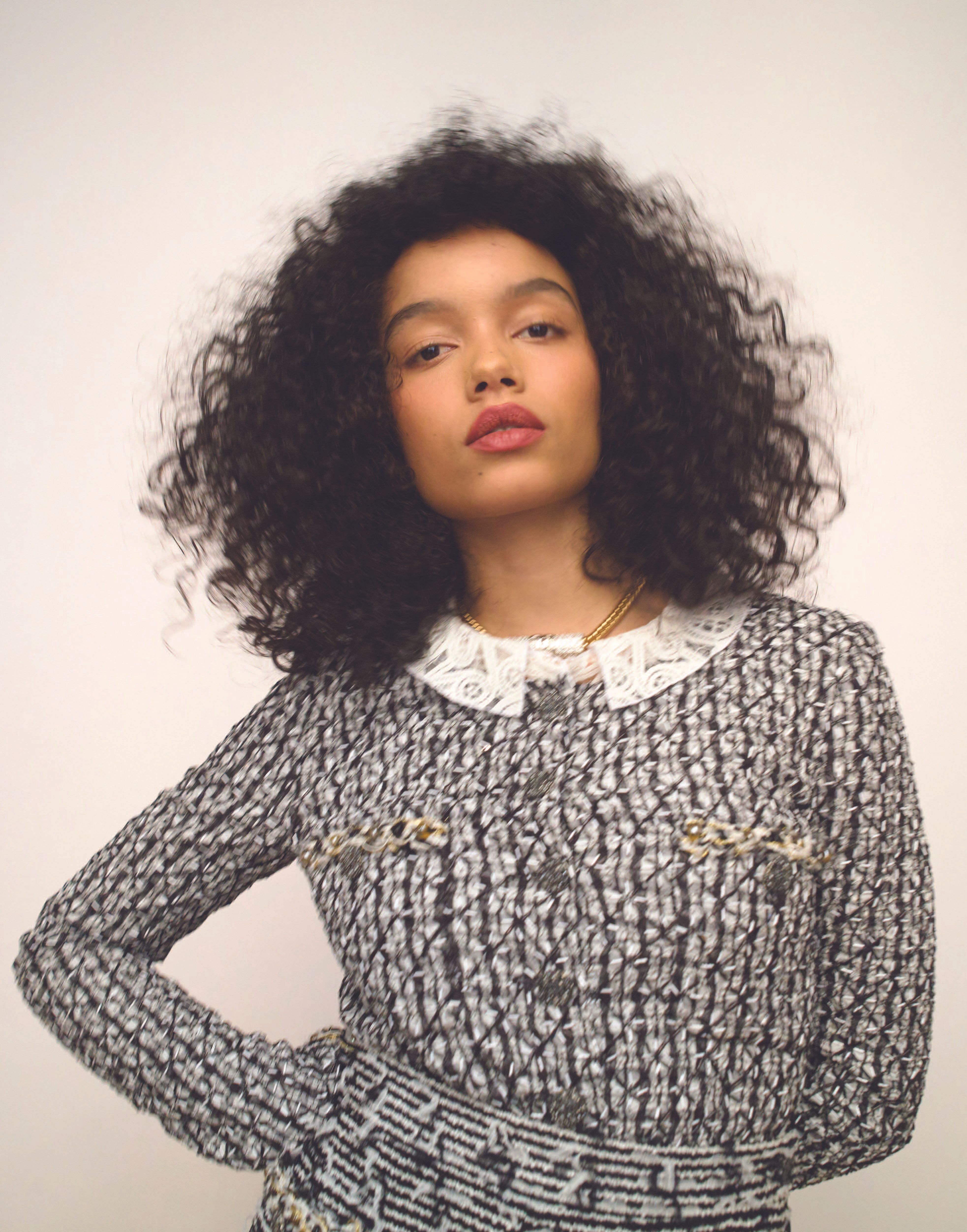 Woman with curly hair poses in a patterned tweed jacket with a lace collar against a plain studio backdrop, fashion editorial shot.