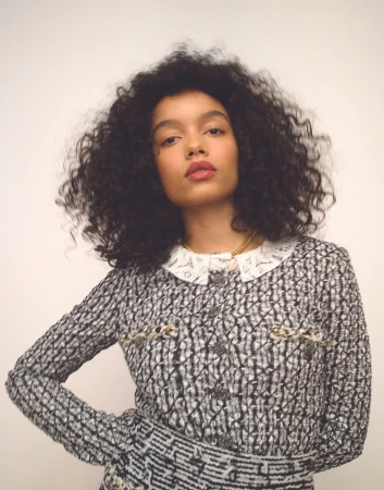 Woman with curly hair poses in a patterned tweed jacket with a lace collar against a plain studio backdrop, fashion editorial shot.
