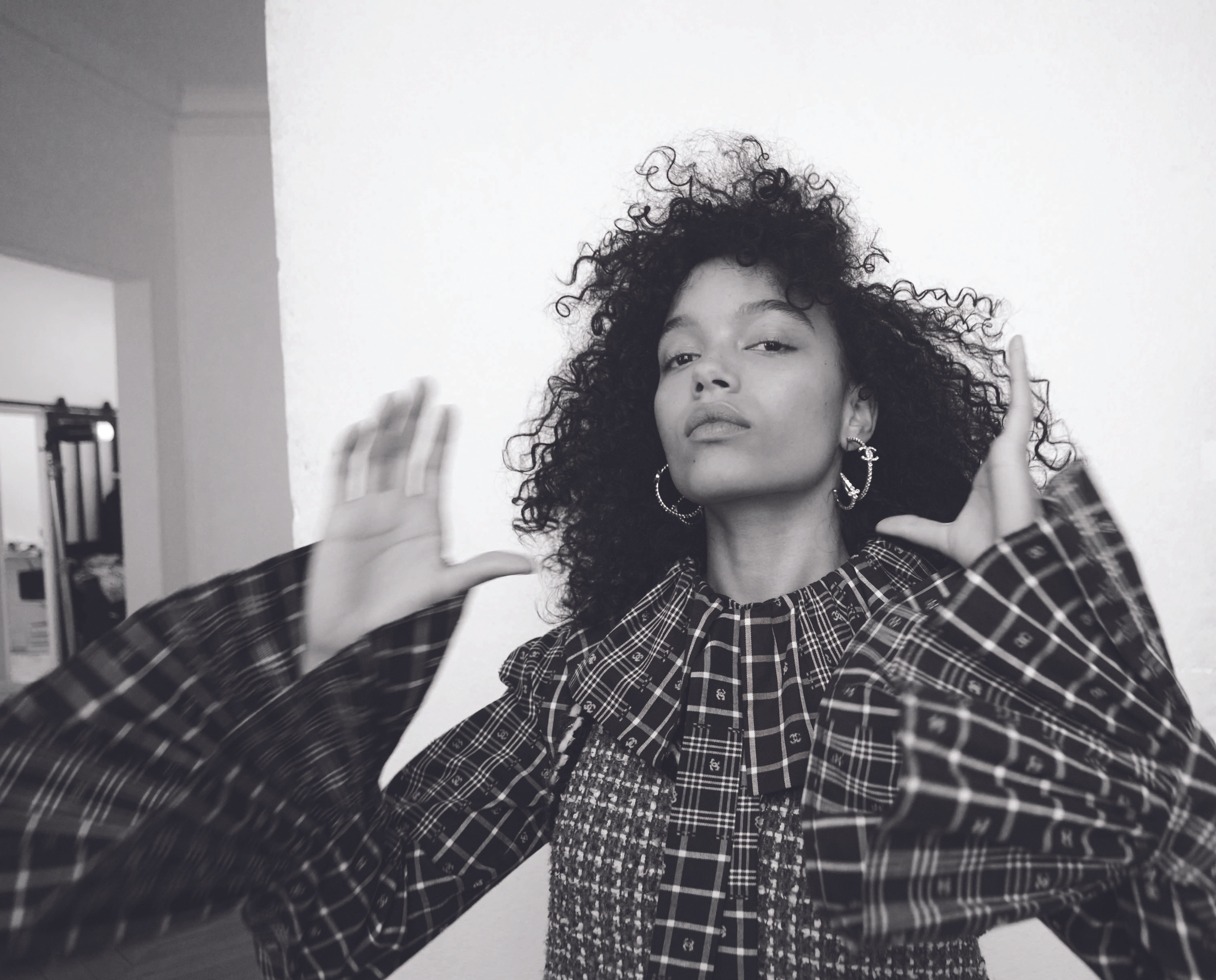 Monochrome portrait of Whitney Peak with dark curly hair wearing a plaid ruffled top and hoop earrings against a white wall.