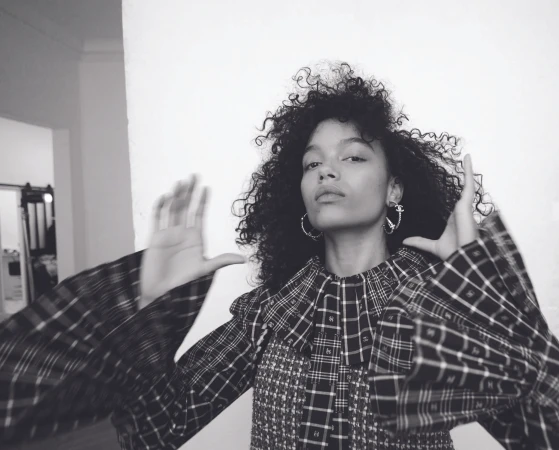 Monochrome portrait of Whitney Peak with dark curly hair wearing a plaid ruffled top and hoop earrings against a white wall.
