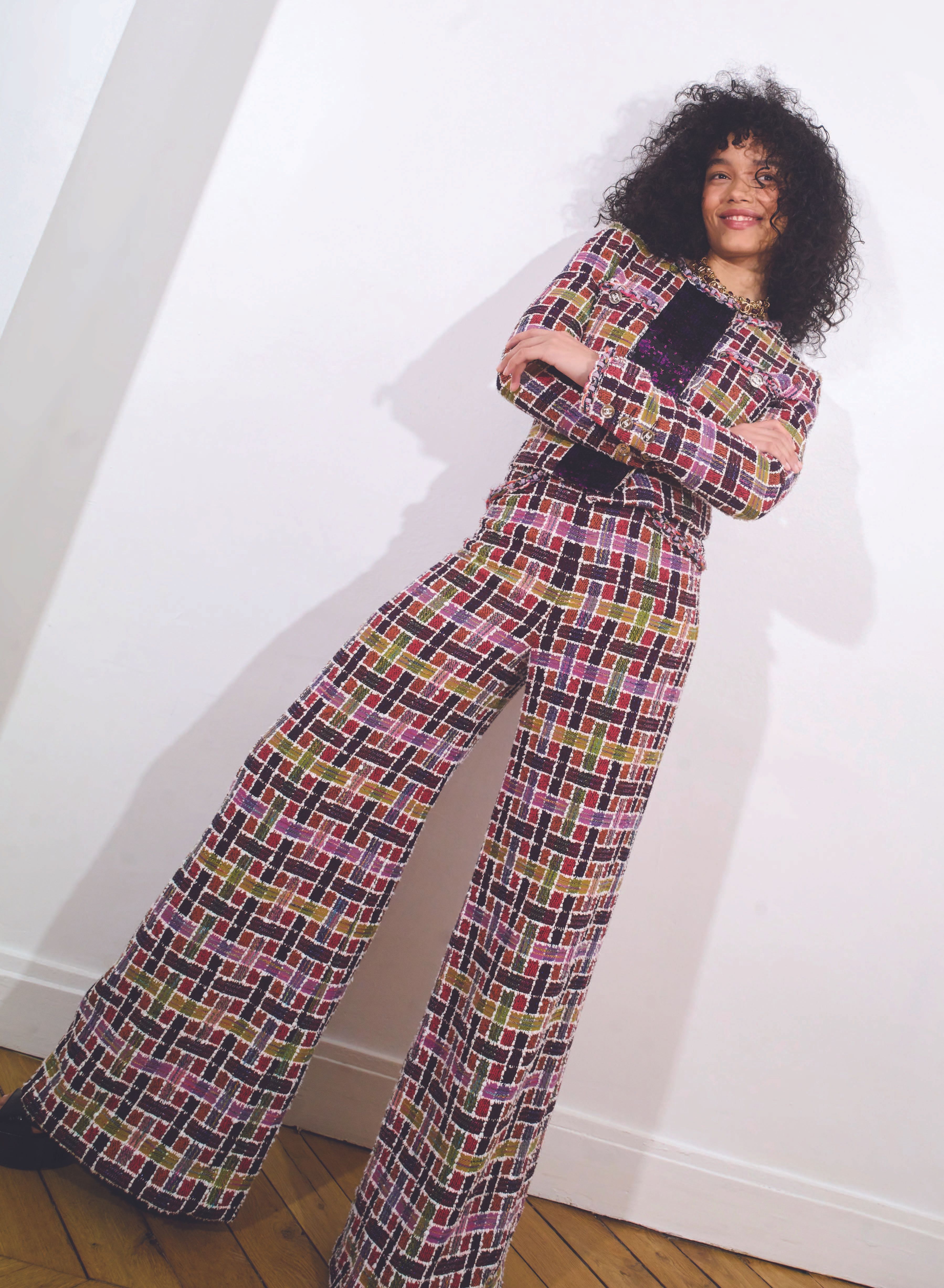 A young adult woman with curly hair poses in a patterned jacket and pants against a white background, arms crossed, smiling in an indoor space.