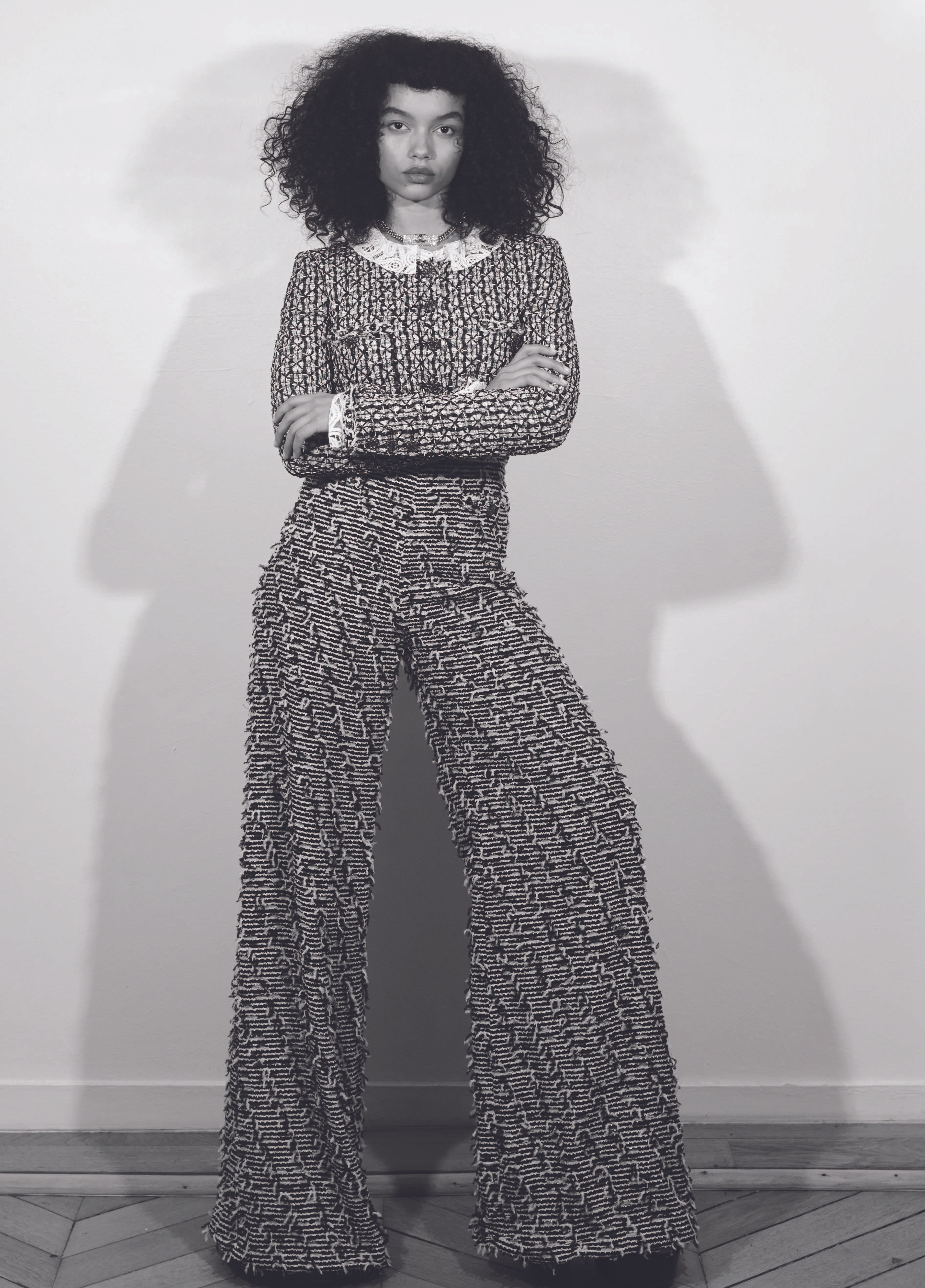 Whitney Peak in a patterned tweed jacket and trousers posing against a plain wall with a sharp shadow cast behind her.