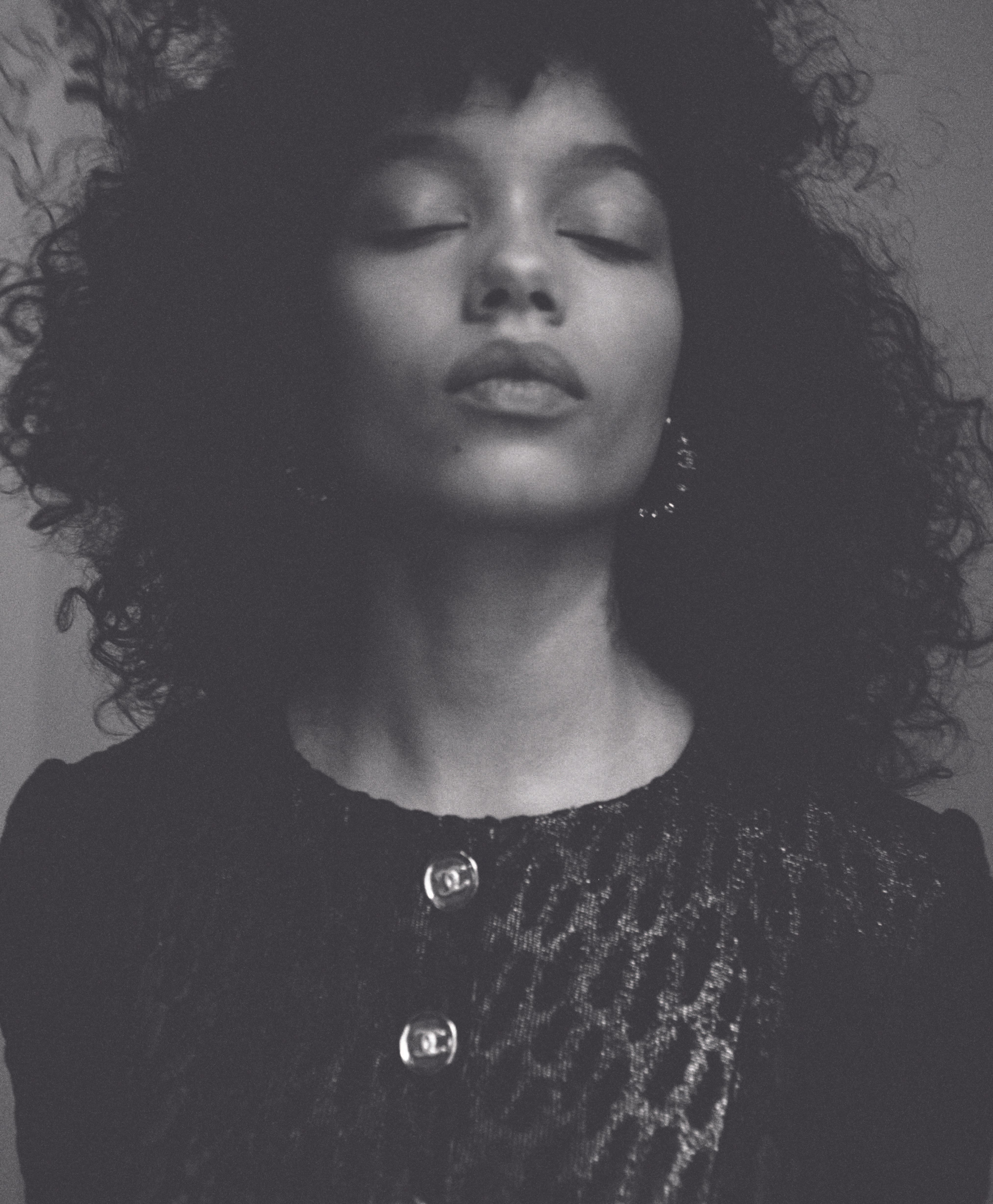 Black and white portrait of a woman with curly hair, eyes closed, wearing a detailed jacket with button embellishments. 