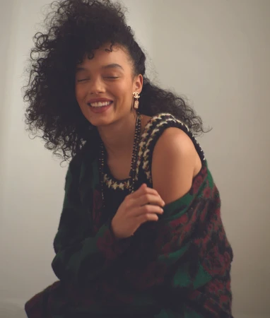 Portrait of Whitney Peak smiling with voluminous curly hair wearing a patterned knit top against a plain light background