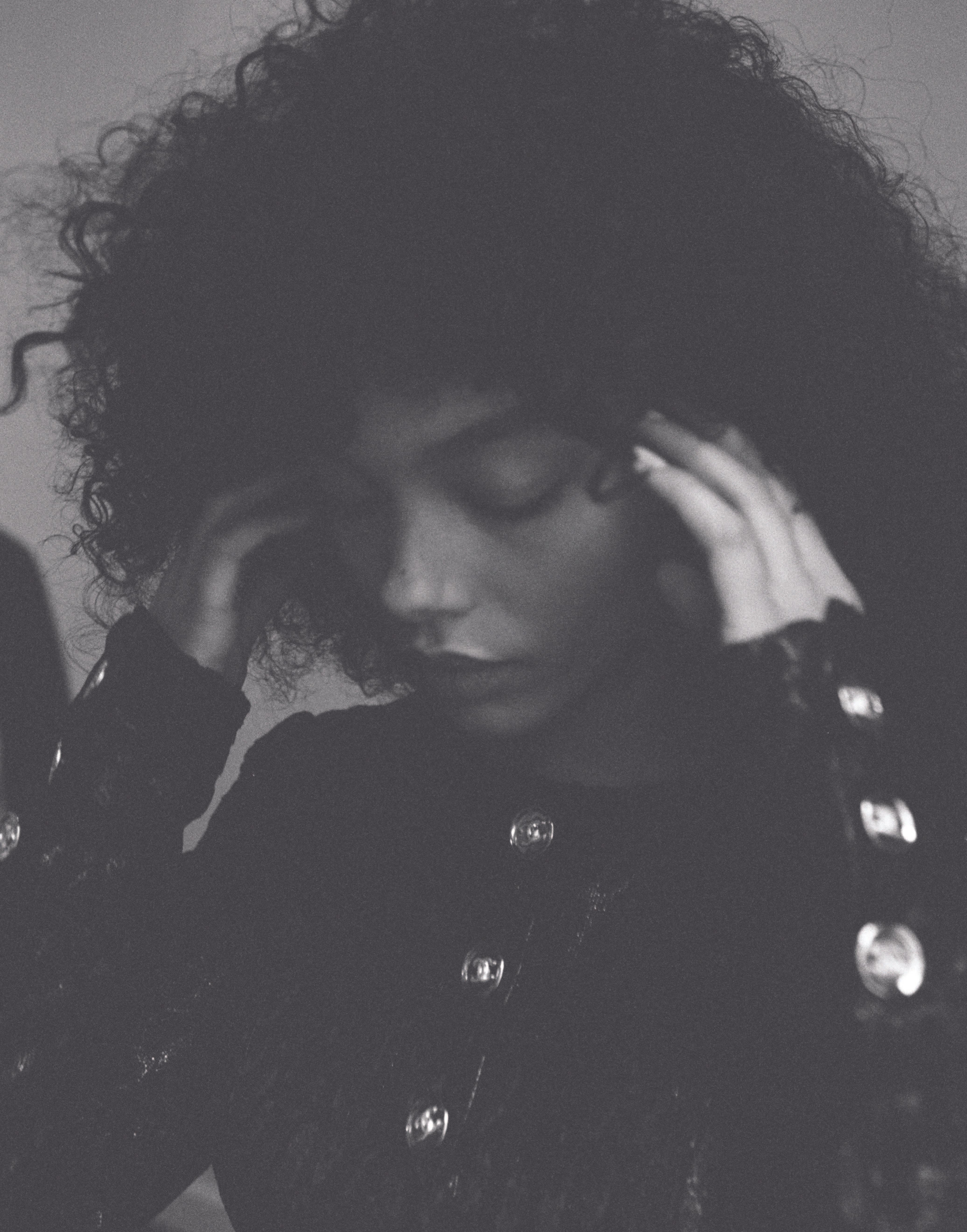 Close-up portrait of Whitney Peak wearing a dark buttoned top with voluminous curly hair, holding hands to temples.