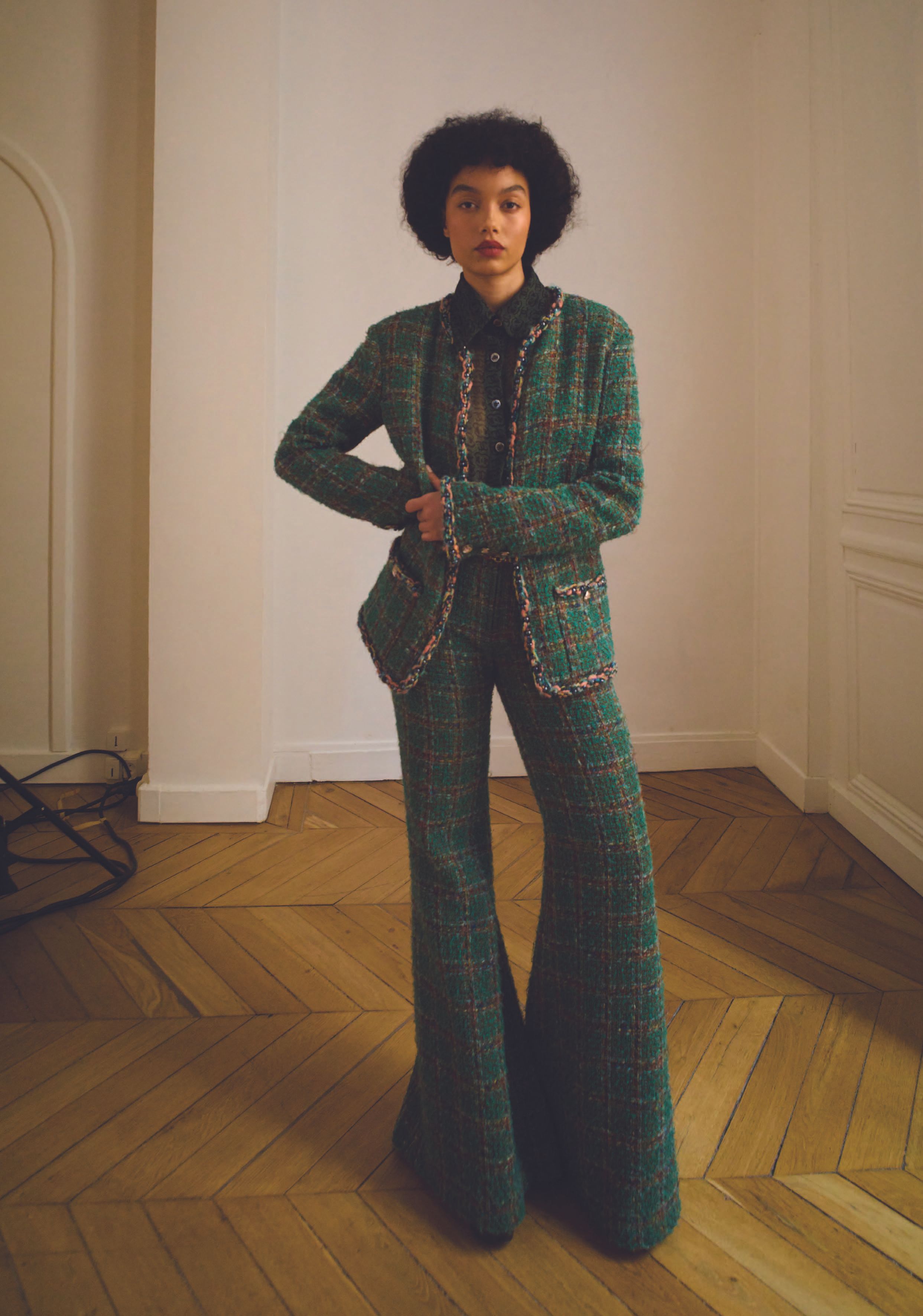 A young adult woman poses indoors wearing a green tweed jacket and matching flared pants in a fashion editorial.