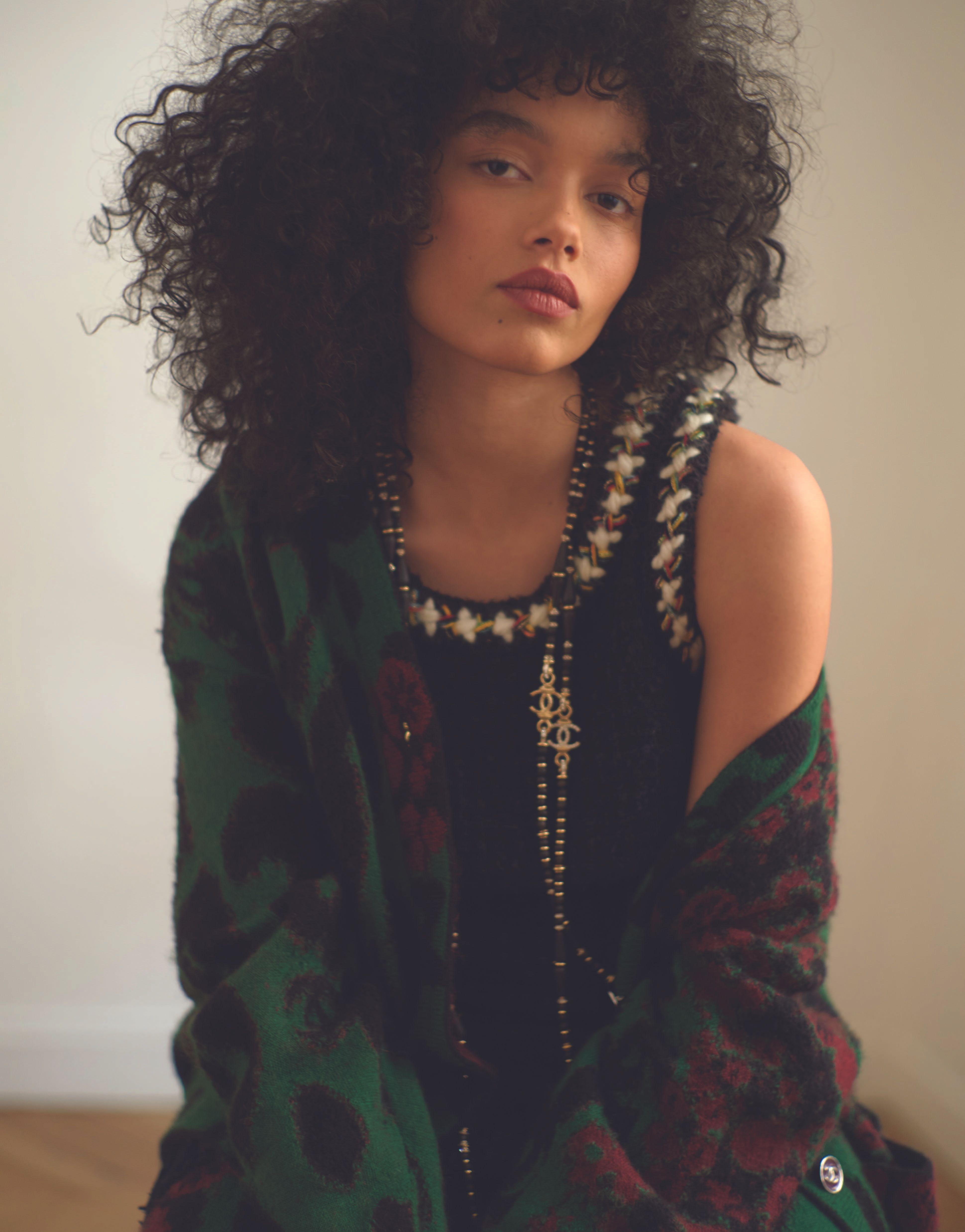 Brunette woman with voluminous curls in a black top with beaded neckline and statement necklace, draped in a green and red patterned cardigan.