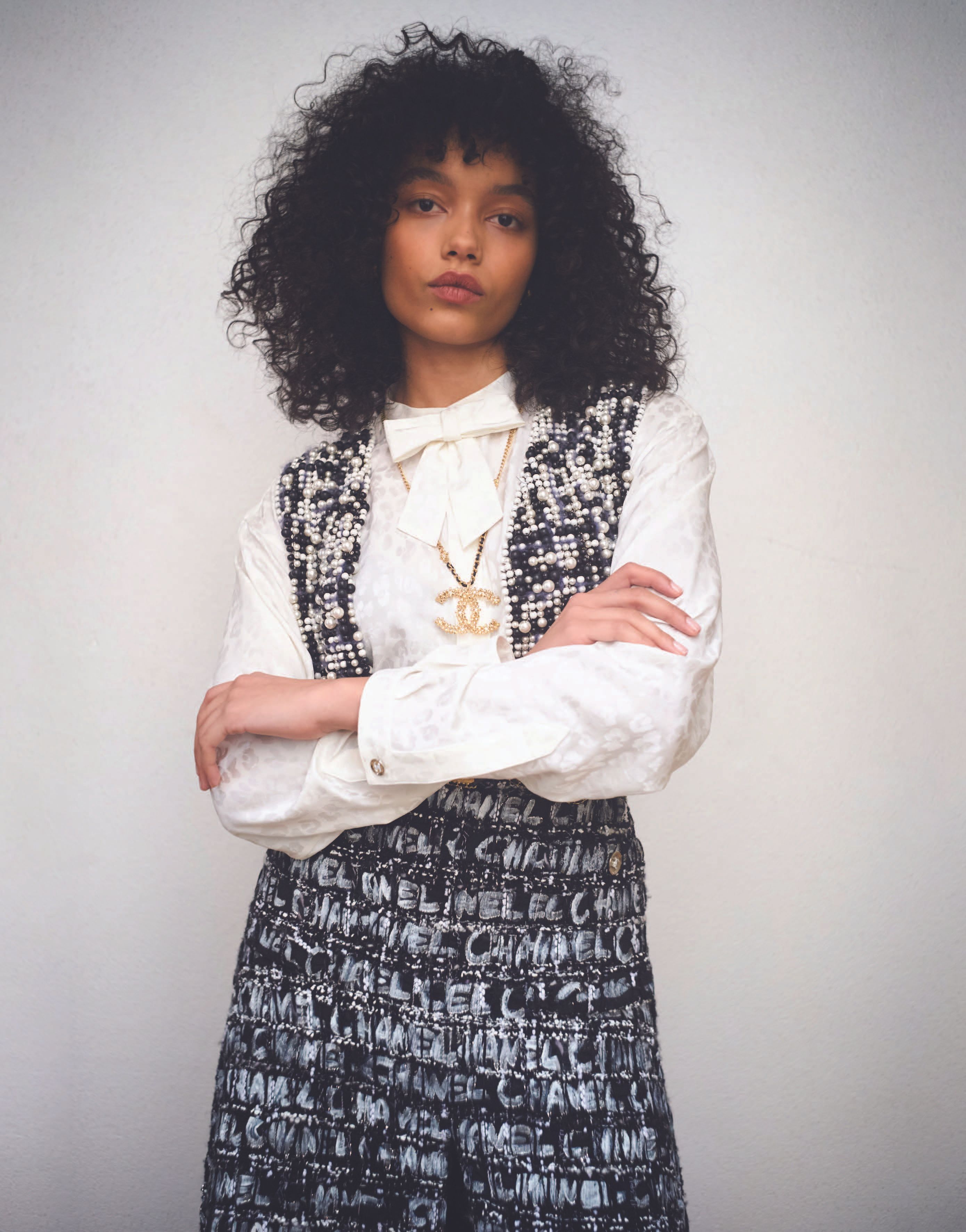 Whitney Peak with dark curly hair wearing a white blouse with bow and patterned tweed vest against a plain background